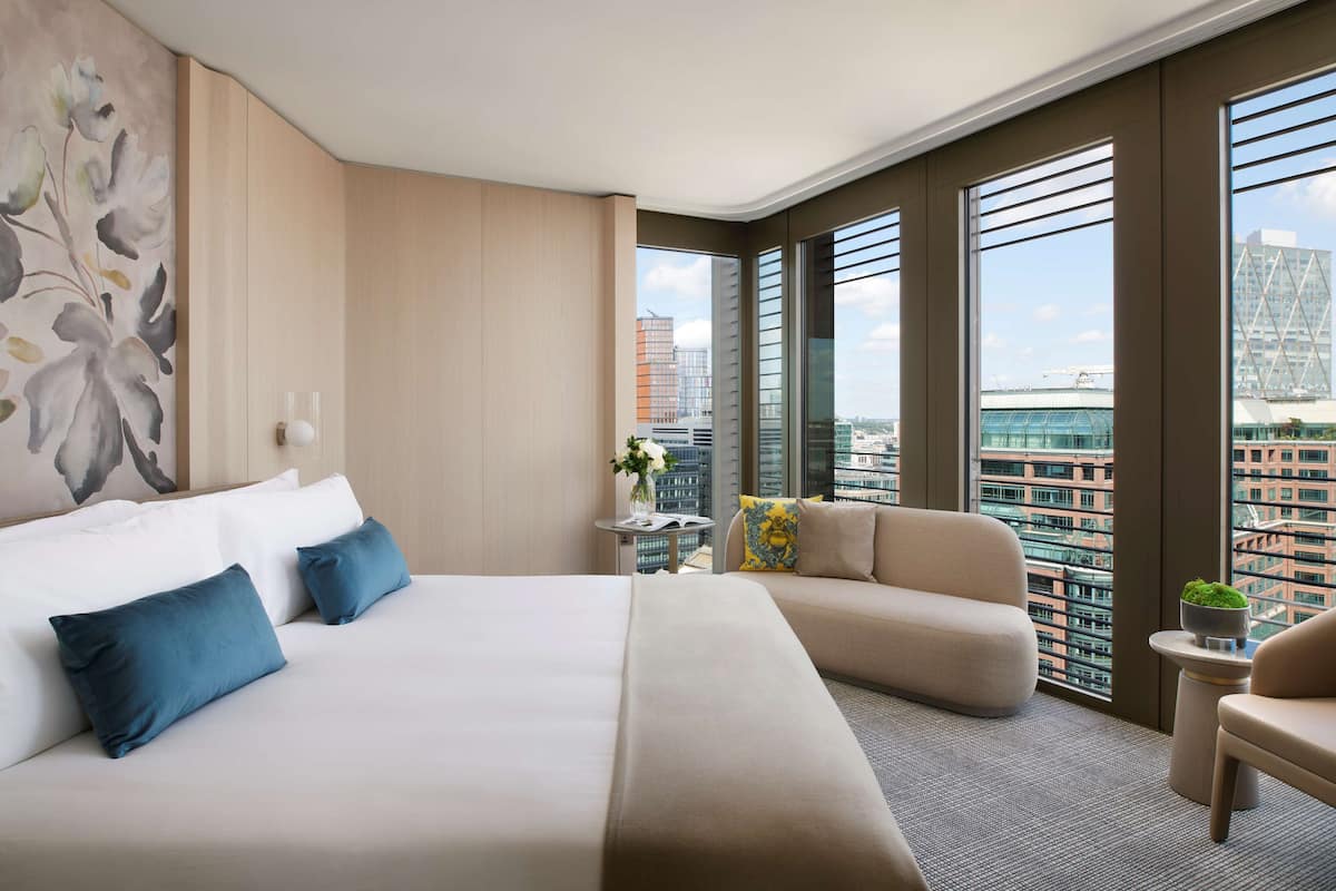 A modern hotel room with a large bed, a sofa, a small table with a plant, and a city view through large windows.