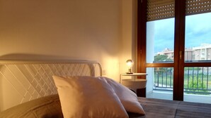 Ein Schlafzimmer mit einem Bett, einem Nachttisch mit Lampe und einem Fenster mit Blick auf Gebäude und Grünflächen.