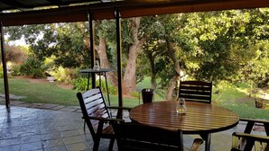Un patio exterior con una mesa redonda de madera y dos sillas, rodeado por un jardín con árboles y plantas.