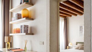 A hotel room with a bed, a desk with a lamp, shelves with books and decorative items, and a view of the outside through a window. - Lion Palace Junior Suite R&R (Venecia)