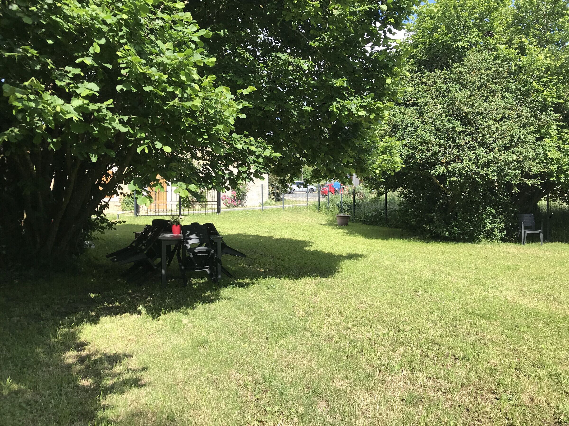 A grassy outdoor area with a picnic table and chairs, surrounded by trees and a fence.