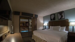 Habitación de hotel con una cama grande, lámparas de noche, un escritorio con lámpara, televisor y puerta que da a un balcón con piscina.
