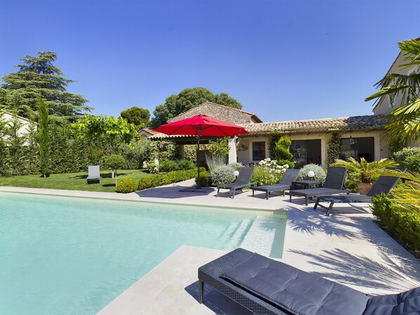 Un espace piscine avec des chaises longues et un parasol rouge, entouré de verdure et bordé d’un bâtiment dont le toit est recouvert de tuiles.