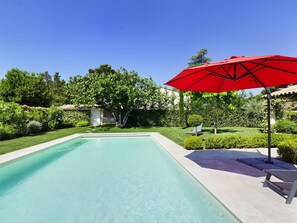 Une piscine avec un parasol rouge, entourée d’une végétation bien entretenue et sous un ciel bleu dégagé.