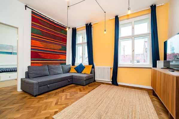 A modern living room with a grey sectional sofa, a colorful abstract wall hanging, and a large window with curtains.