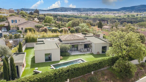 A modern house with a pool, surrounded by lush greenery and a scenic view of the sea and hills.