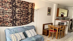 Un salon comprenant un canapé, une table basse en bois, une table à manger avec des chaises, un coin cuisine avec un four à micro-ondes et une tapisserie à motifs accrochée au mur.