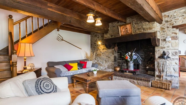 Una sala rústica con chimenea de piedra, vigas de madera, un sofá con cojines coloridos y una escalera.