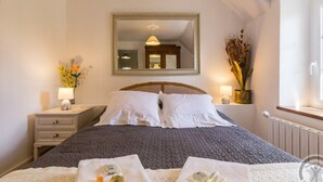 Una cama bien hecha con colcha gris, una mesita de noche con lámpara, un espejo sobre la cama y una ventana con vistas al exterior.