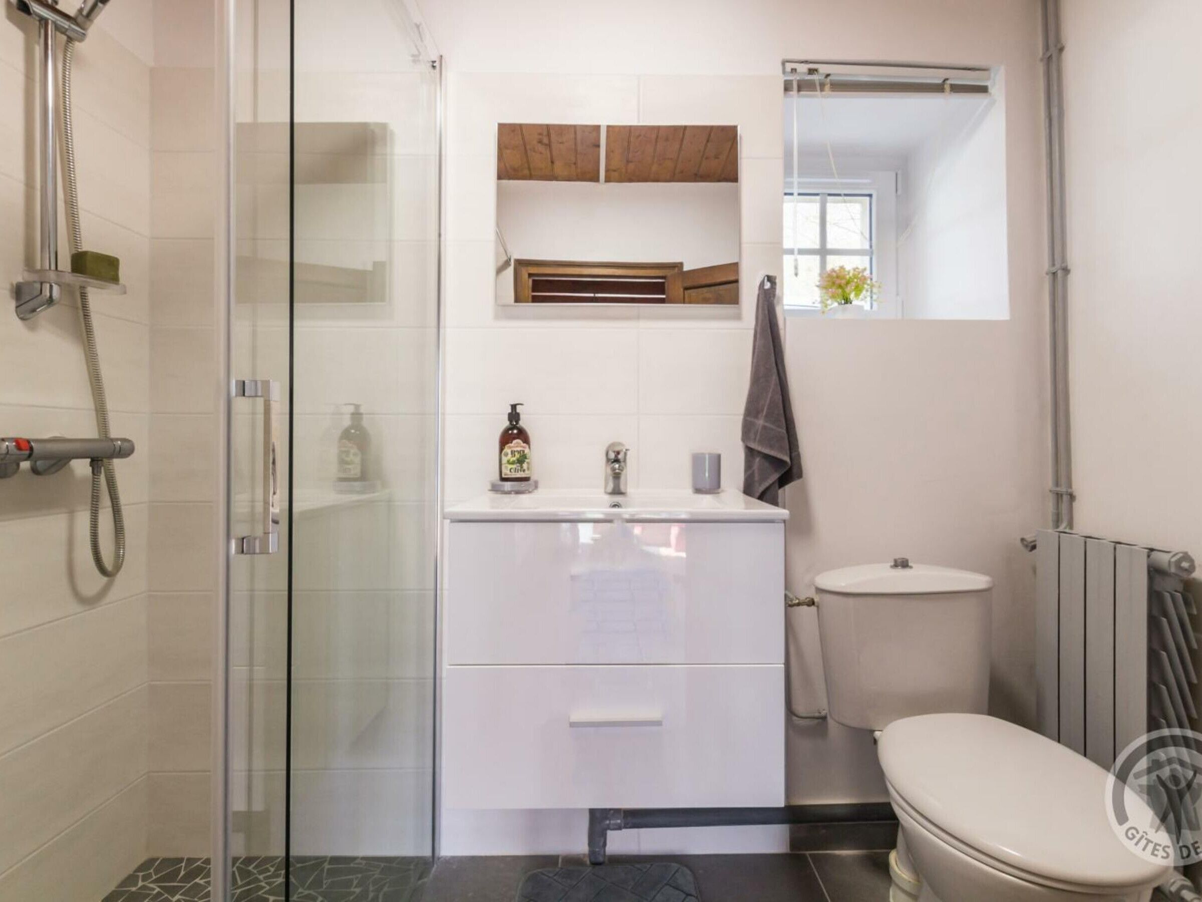 Une salle de bain moderne avec une douche à parois de verre, un lavabo blanc doté d’un distributeur de savon, des toilettes et une petite fenêtre située au-dessus du miroir.