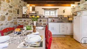 Una cocina rústica con paredes de piedra, vigas de madera, armarios blancos y un comedor listo para una comida.