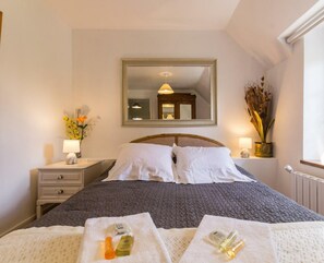 A neatly made bed with a grey quilt, a bedside table with a lamp, a mirror above the bed, and a window with a view of the outside.