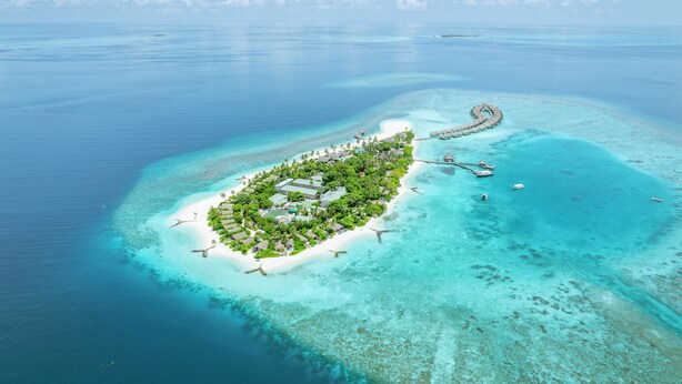 An aerial view of a tropical island resort with clear turquoise waters, a white sandy beach, and overwater bungalows connected by a curving walkway. - Cora Cora Maldives-Premium All-Inclusive (Maamigili Island)
