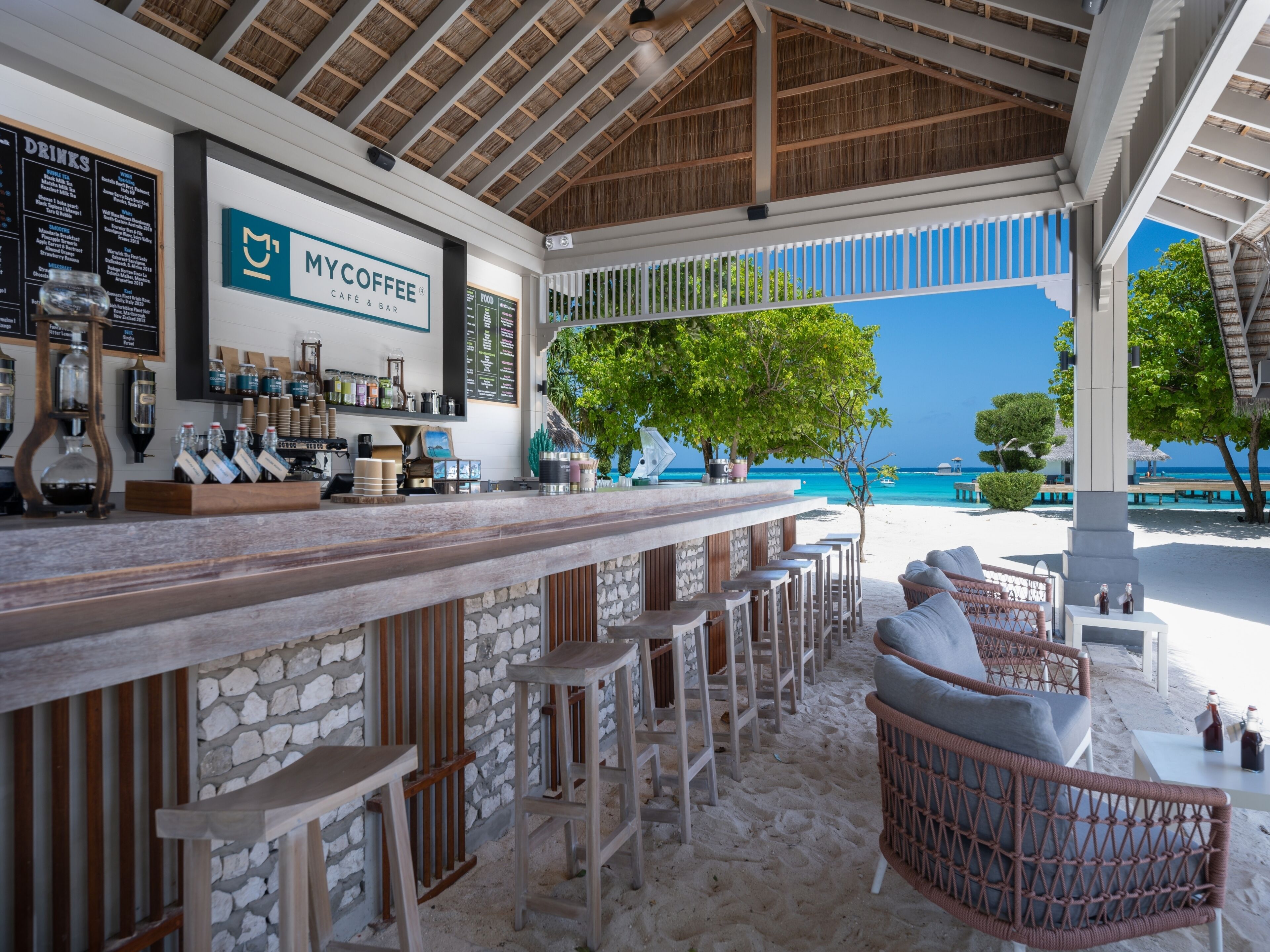 Un bar frente a la playa con mostrador de piedra, taburetes y mobiliario de mimbre.