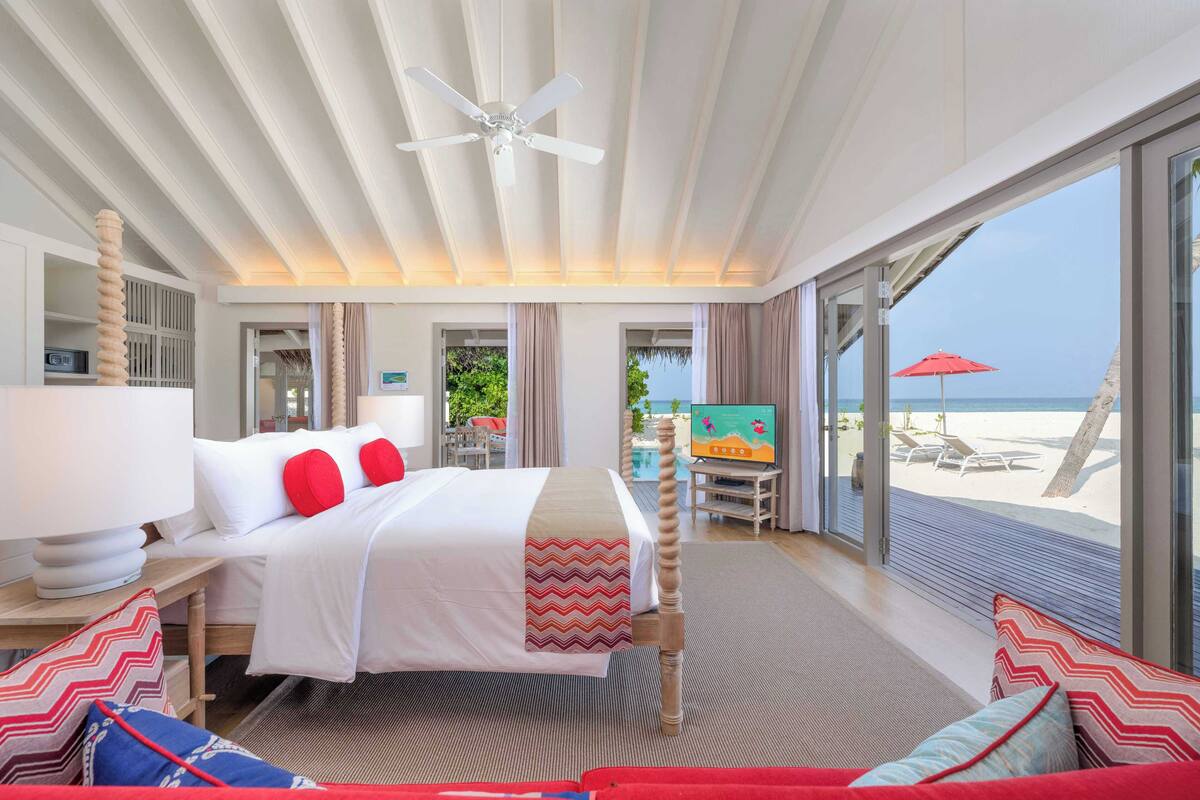 A hotel room with a bed, a TV, a desk, and a view of the beach.