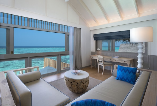 A modern room with a large window overlooking the ocean, a round stone table, a sofa, a desk, and a lamp. - Cora Cora Maldives-Premium All-Inclusive (Maamigili Island)