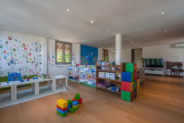A modern, well-lit room with a whiteboard filled with children's handprints, a white table with toys, a bookshelf with books and toys, and a TV in the background. - Cora Cora Maldives-Premium All-Inclusive (Maamigili Island)