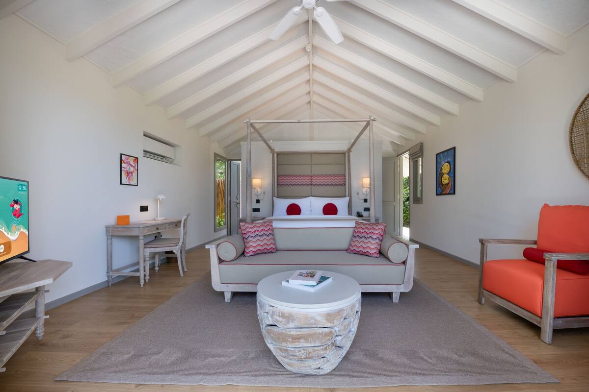 A room with a high ceiling, a four-poster bed, a sofa, a coffee table, a desk, and a television.