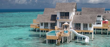 Overwater bungalows with thatched roofs, a pool, and wooden walkways over clear turquoise water.