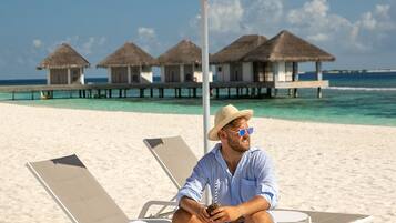 Eine Strandansicht mit einer Person, die auf einer Liege unter einem roten Sonnenschirm sitzt, einer Strandtasche und Overwater-Bungalows im Hintergrund.