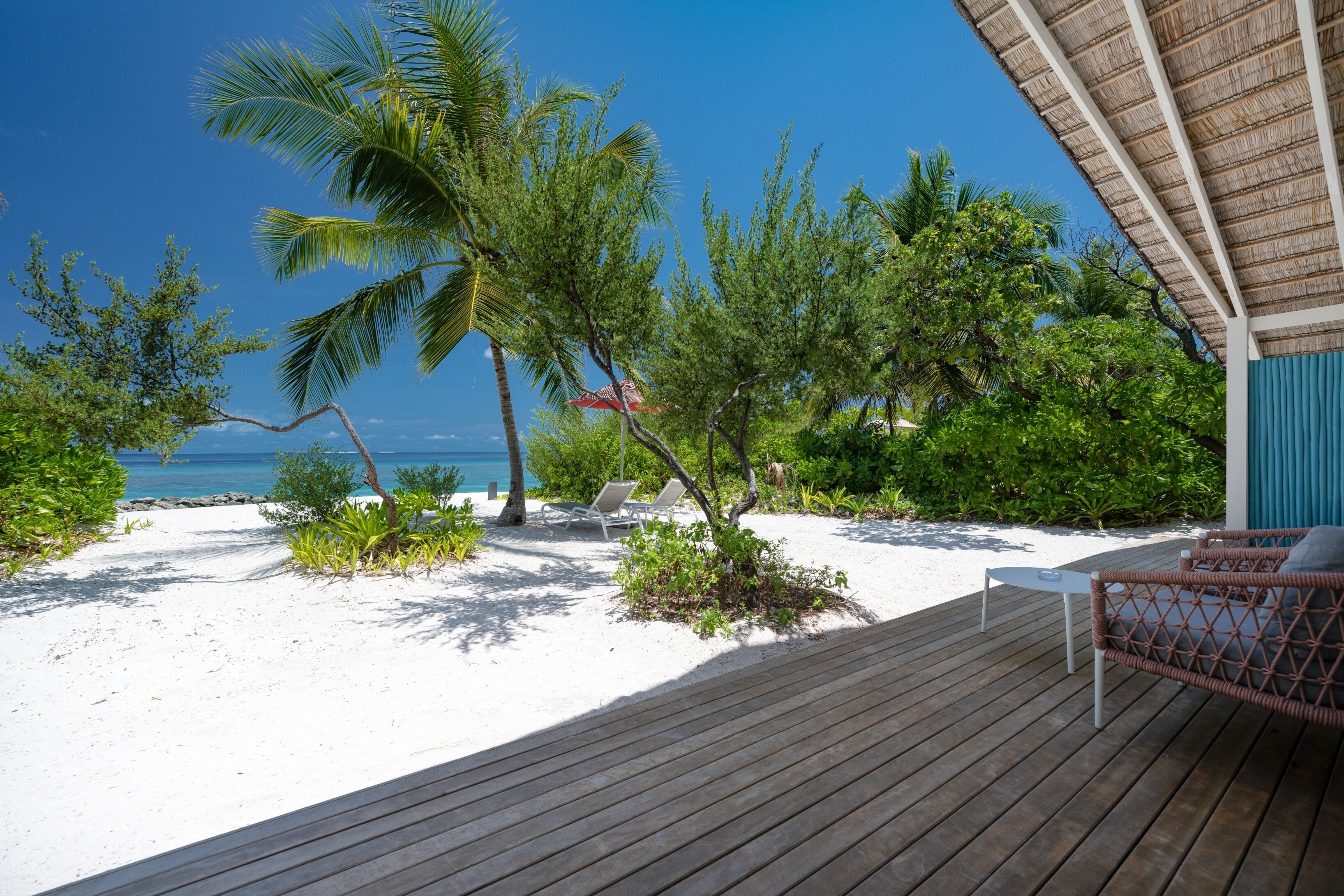 A wooden deck with lounge chairs and a table, overlooking a beach with palm trees and a clear blue sky.