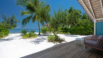 A wooden deck with lounge chairs and a table, overlooking a beach with palm trees and a clear blue sky.