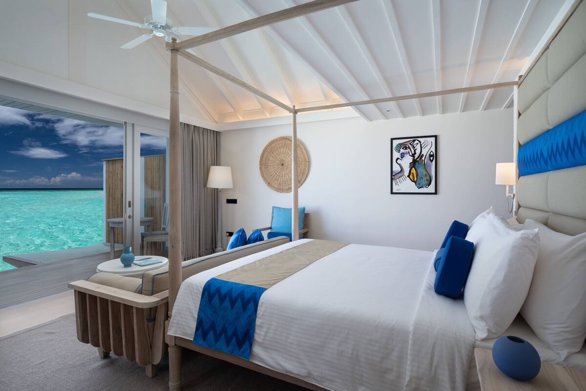A four-poster bed with a view of the ocean, a round woven wall art, and a blue pillow on the bed.