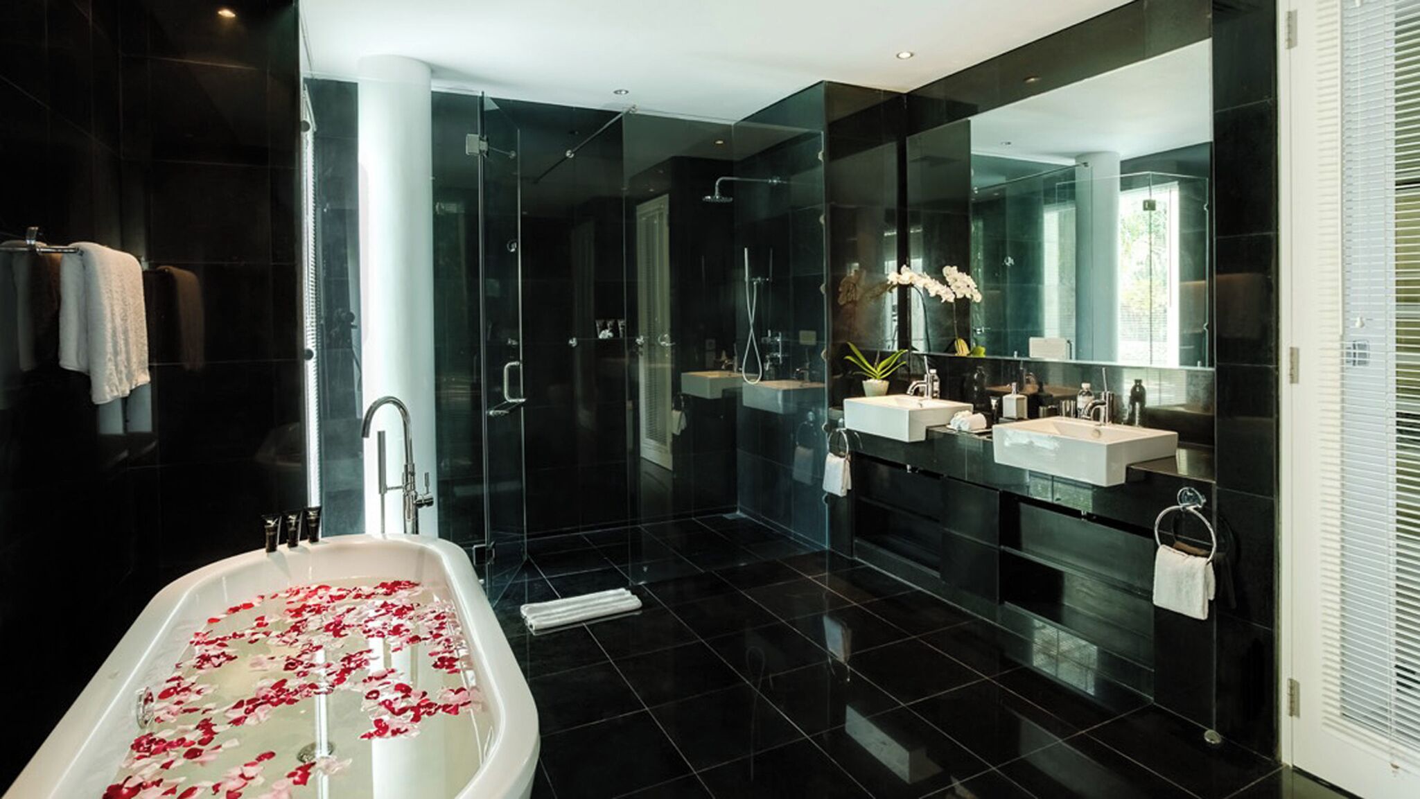 A modern bathroom with a glass-enclosed shower, a freestanding bathtub with rose petals, and a large mirror reflecting the room.