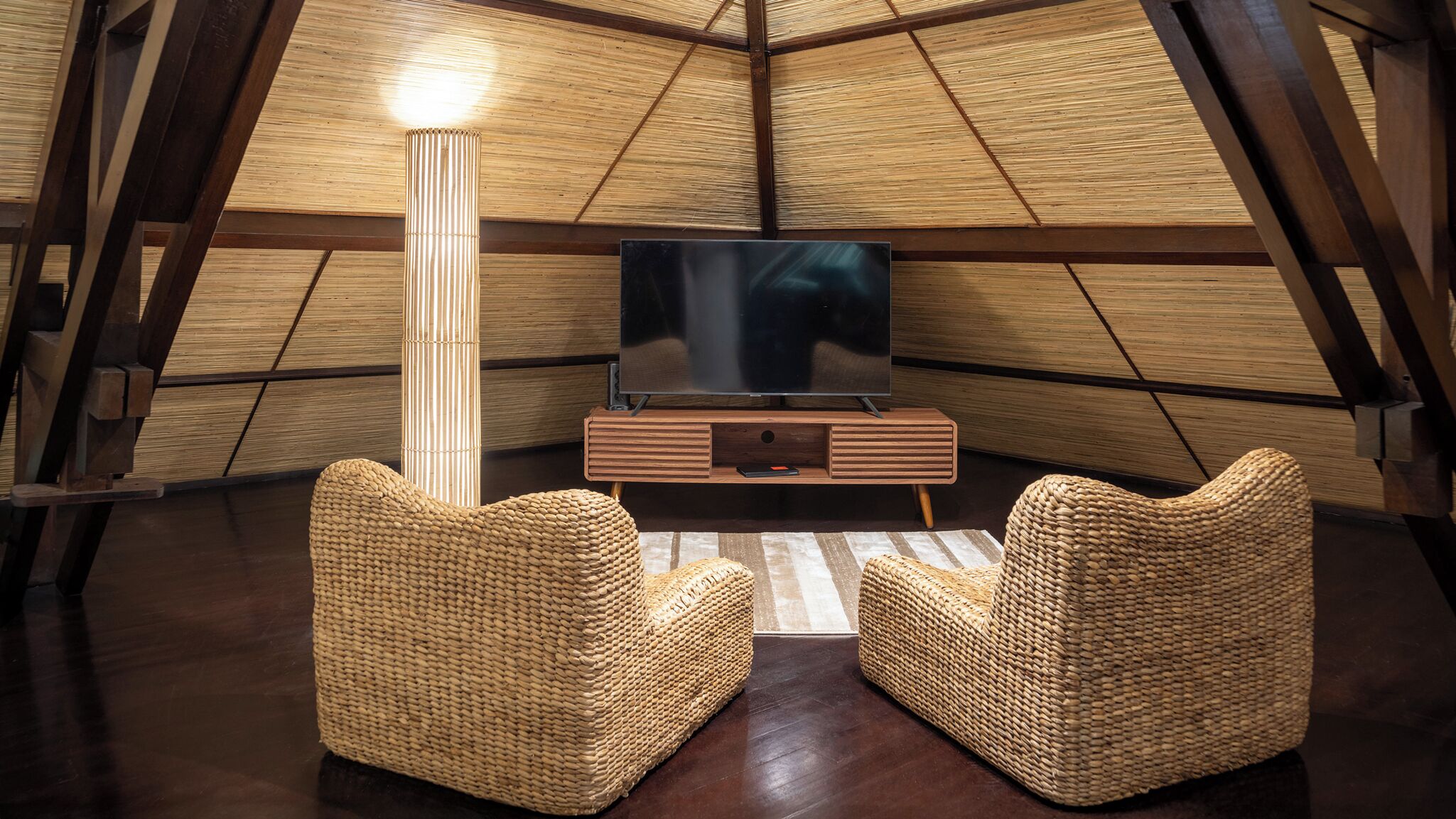 A cozy room with a large flat-screen TV on a wooden stand, two woven chairs, and a tall floor lamp.