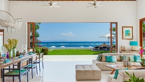 Ein geräumiges Wohnzimmer mit Meerblick, eine Essecke mit Holztisch und -stühlen sowie eine Sitzecke mit Sofa und Hocker.