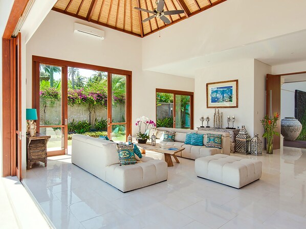 A spacious living room with a high ceiling, large windows, and a view of a lush garden. - Villa Tirta Nila by Elite Havens (Manggis)
