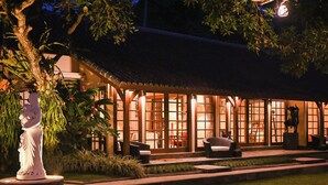 A traditional Japanese-style building with a thatched roof, illuminated windows, and a statue in the garden. - Villa San (Ubud)