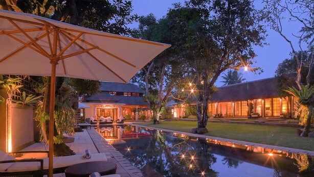 A poolside area with lounge chairs and a table, a reflection pool, and a building with warm lighting. - Villa San (Ubud)