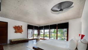 Una camera da letto moderna con un letto grande, una panca, una ventola a soffitto e vista su un giardino attraverso ampie finestre.