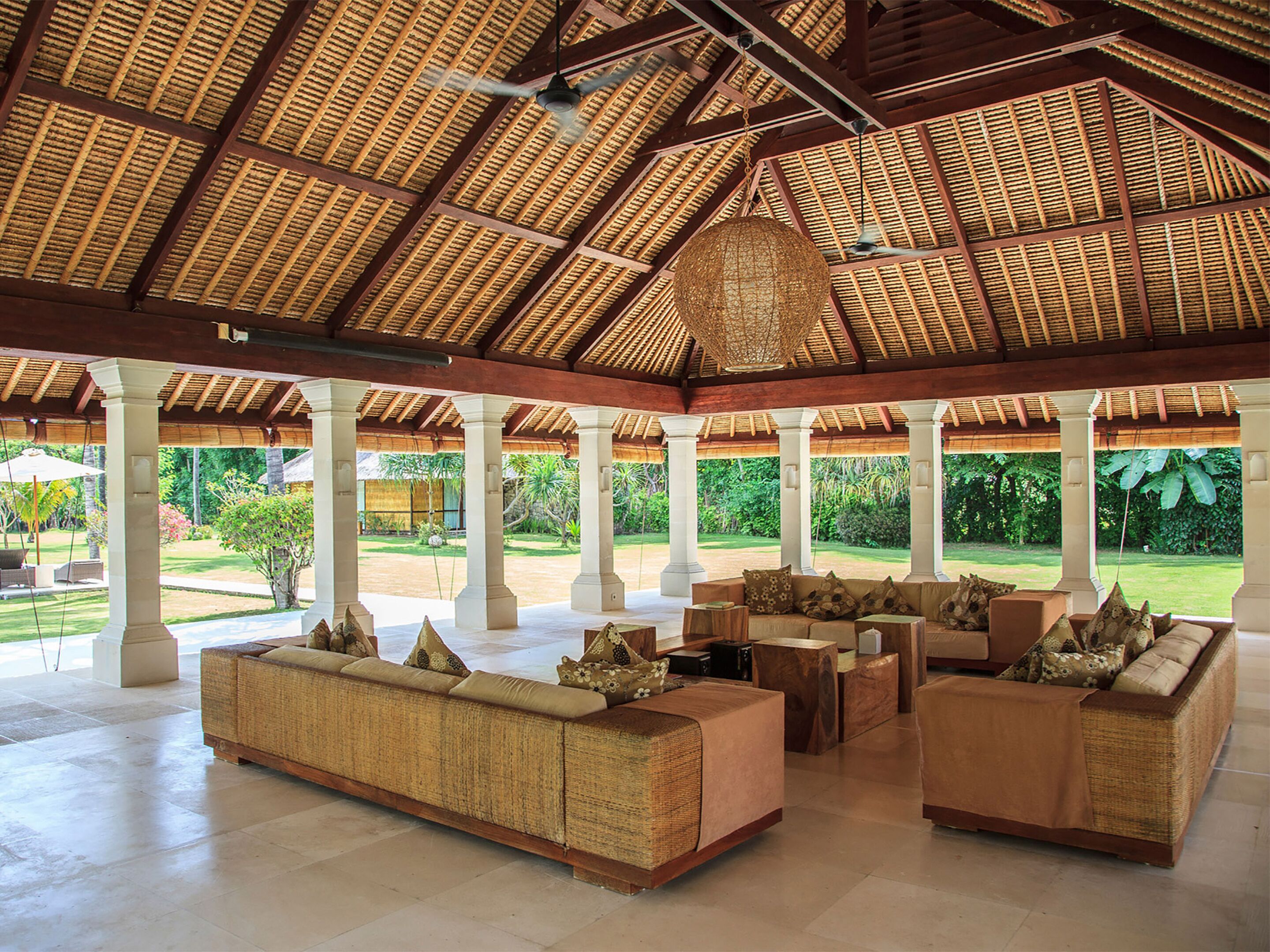 A spacious covered lounge area with a high thatched roof, multiple seating arrangements, and a view of a lush garden.