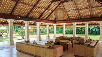 A spacious covered lounge area with a high thatched roof, multiple seating arrangements, and a view of a lush garden.
