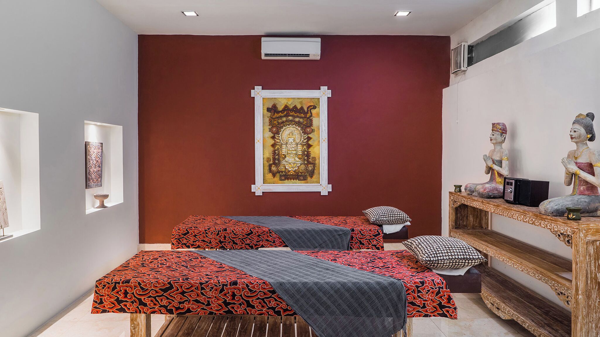 A room with two beds, a wall-mounted air conditioner, a framed artwork, and a shelf with statues.