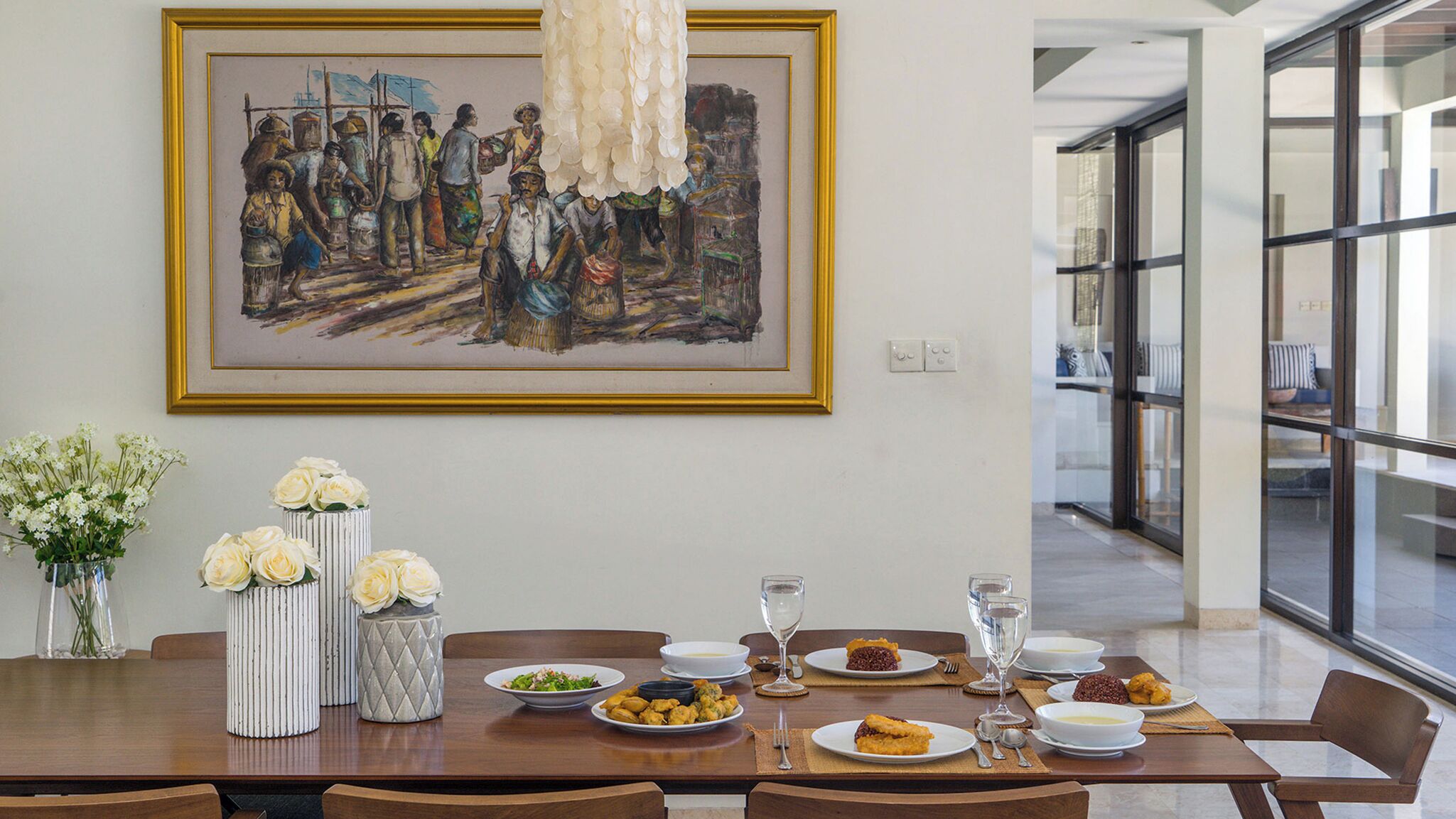 A dining area with a large painting, white vases with flowers, and a table set for a meal.
