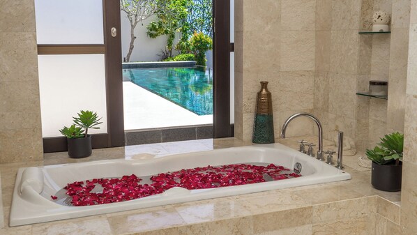 A bathroom with a bathtub filled with rose petals, a glass shower enclosure, and a view of a swimming pool. - Villa Adenium by Elite Havens (Jimbaran)