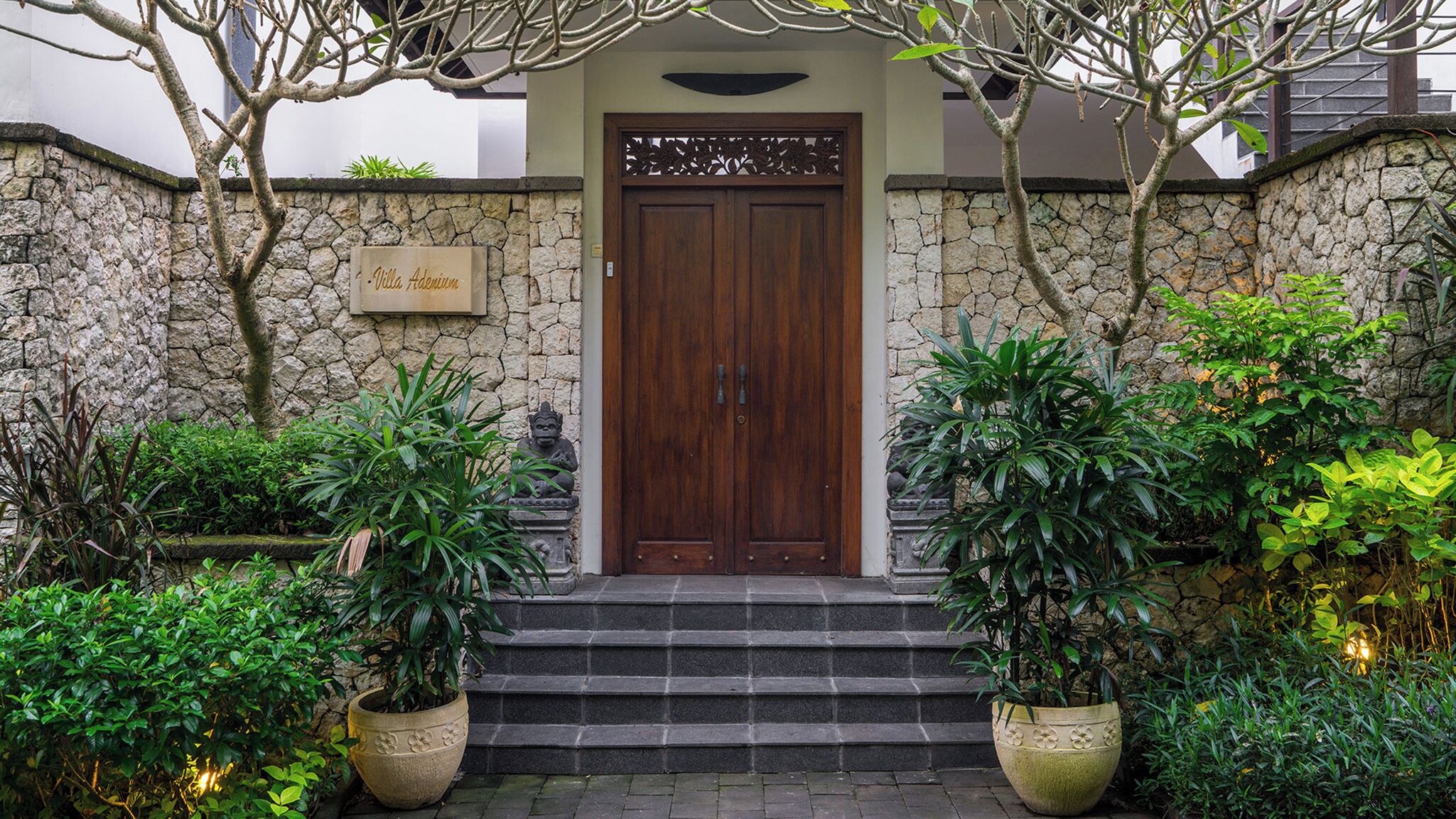 Uma entrada com paredes de pedra, uma porta de madeira, plantas em vasos e uma placa com a inscrição 'Villa Adenium'.
