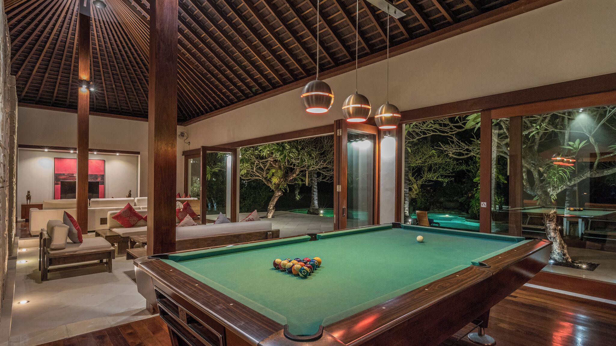 Un espace piscine comprenant une table de billard verte, des fauteuils confortables et une vue sur un jardin, grâce à de grandes portes vitrées.