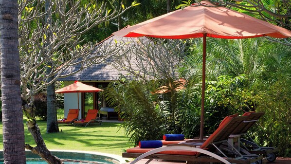 Zona de piscina con sillas de descanso y sombrillas, rodeada de exuberante vegetación y un pequeño edificio con tejado de tejas.