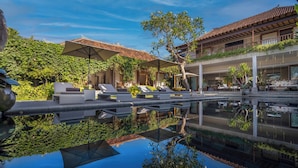 A luxurious outdoor pool area with lounge chairs and umbrellas, surrounded by lush greenery and a traditional building with a tiled roof. - Mandalay Villas (Munggu)