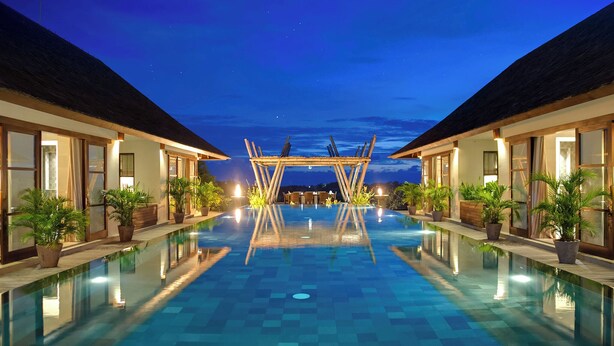 A swimming pool with a wooden deck in the center, flanked by two buildings with large windows and potted plants. - Mandalay Villas (Munggu)