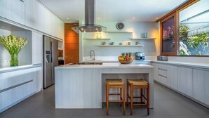 Una cocina moderna con una isla grande, taburetes de madera, estufa empotrada y un amplio ventanal con vistas a la vegetación.