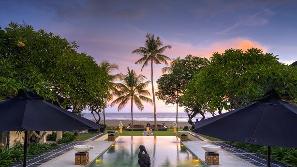 A poolside area with lounge chairs and umbrellas, a statue, and a view of the ocean at sunset. - The Ylang Ylang (Blahbatuh)