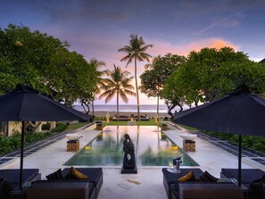 Ein Poolbereich mit Liegestühlen und Sonnenschirmen, einer Statue sowie Blick auf das Meer bei Sonnenuntergang.