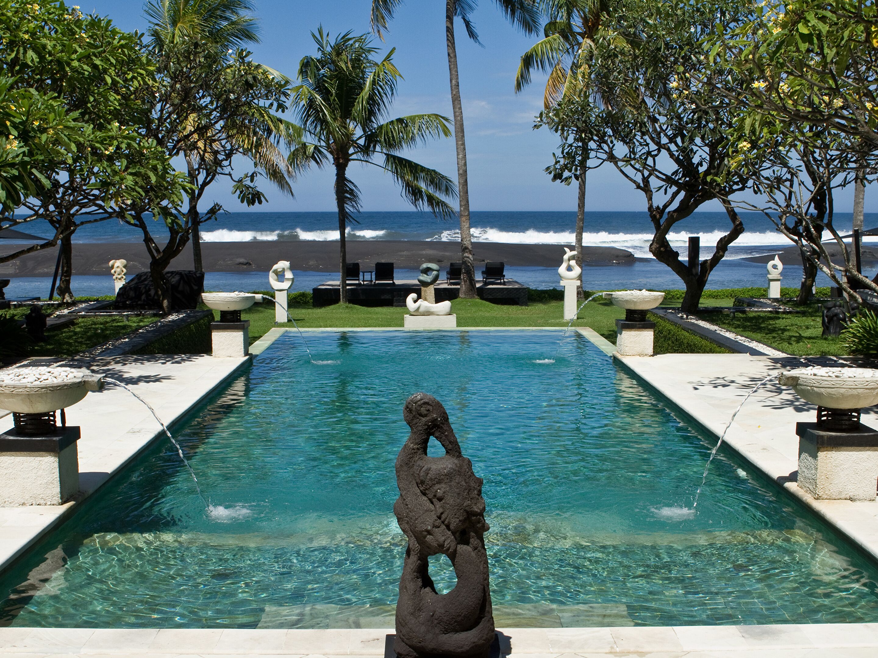 Una piscina con una escultura decorativa en el centro, rodeada de palmeras y vista al océano.