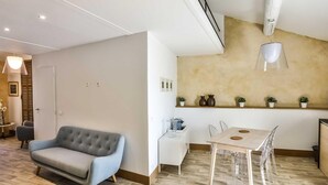 Ein modernes Wohnzimmer mit grauem Sofa, ein Essbereich mit Holztisch und Stühlen sowie eine Küche mit weißem Arbeitsplatz und Topfpflanzen.