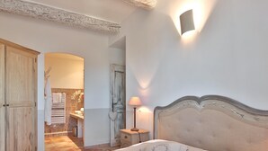 A bedroom with a large bed, a nightstand with a lamp, a wooden wardrobe, and a bathroom visible through an open door. - Apartement Enjoy the Experience Gordes (Gordes)